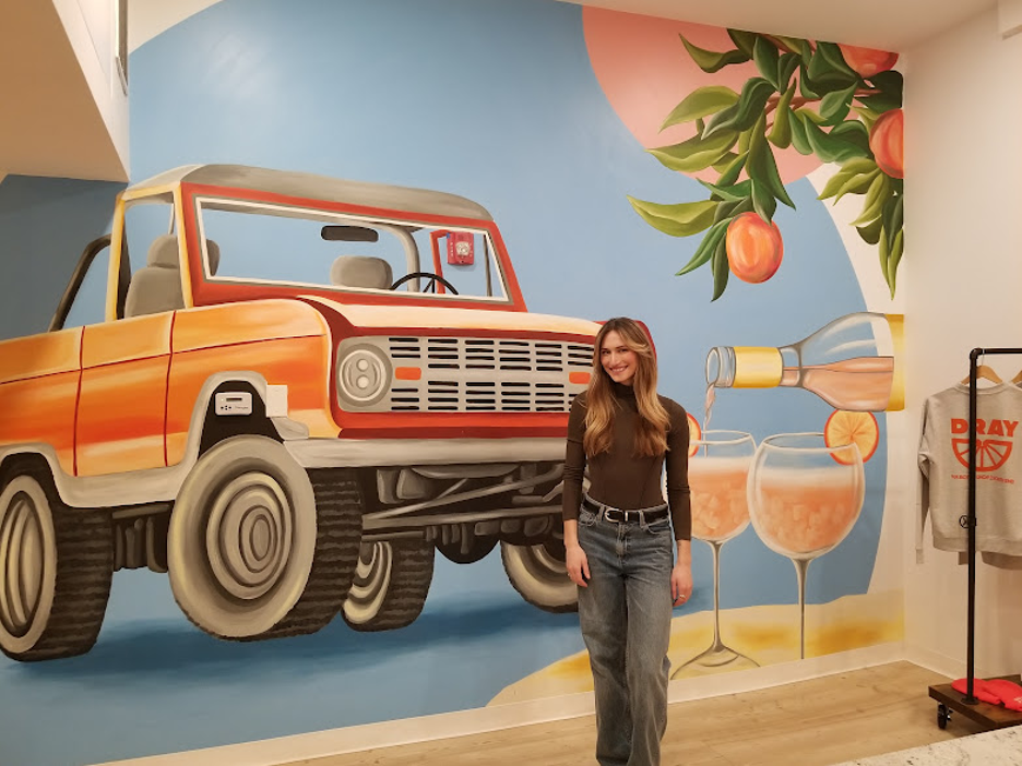 The vintage, orange Ford Bronco mural at Dray, with artist Liv Mileika in front of it.